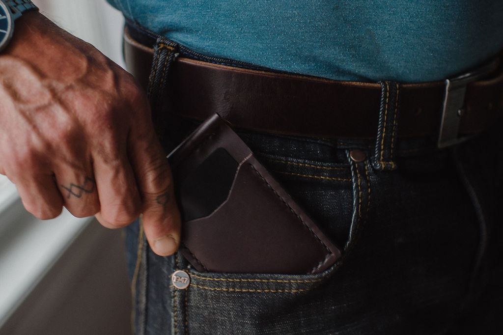 A close-up of a person's hand inserting a dark brown leather card holder or wallet into the back pocket of dark blue jeans, worn with a brown leather belt and a blue shirt.