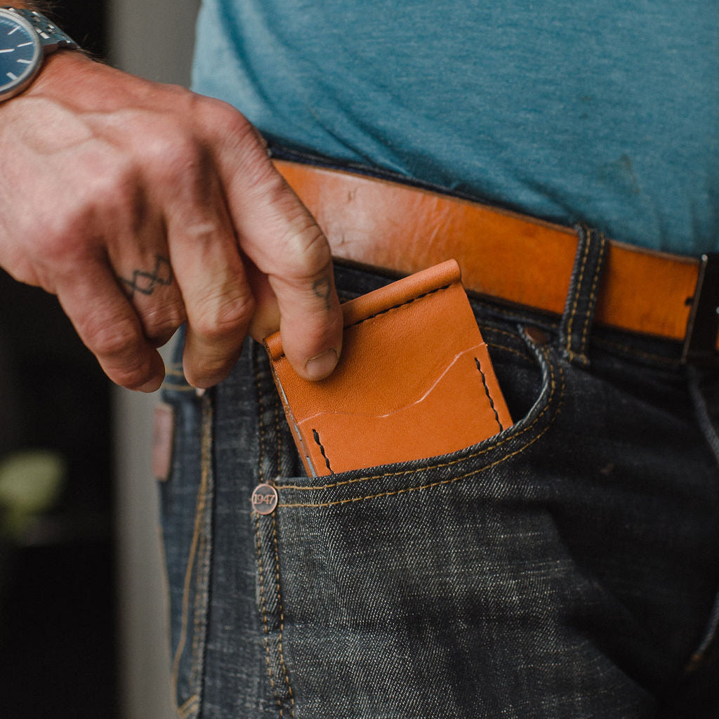 A close-up of a person's hand sliding a slim, tan brown leather card holder or wallet into the pocket of dark blue denim jeans, paired with a tan leather belt.