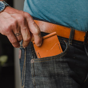 A close-up of a person's hand sliding a slim, tan brown leather card holder or wallet into the pocket of dark blue denim jeans, paired with a tan leather belt.