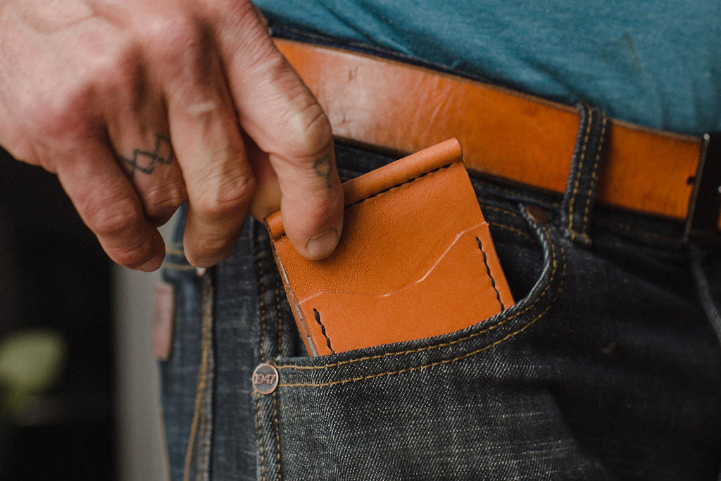 A close-up of a person's hand sliding a slim, light brown leather card holder wallet into the pocket of dark blue denim jeans, paired with a tan leather belt.