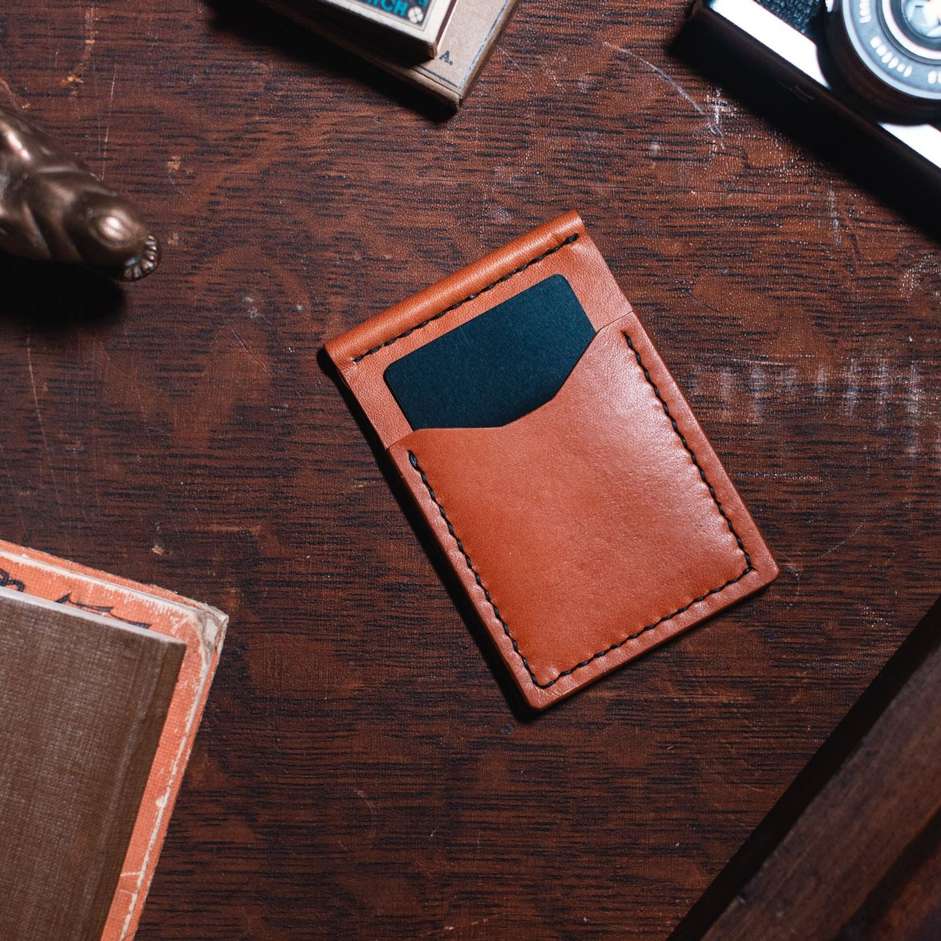A full grain leather card holder wallet in tan colour with a dark blue card inserted, displayed on a dark wooden desk surrounded by vintage props including a camera, a brass alligator figurine, and a book.