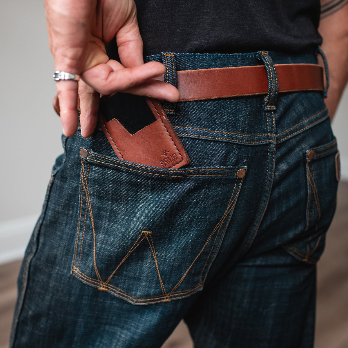 A close-up lifestyle shot of a person wearing dark blue denim jeans with a cognac  brown leather belt and a matching leather cardholder wallet in the back pocket, shown from behind.