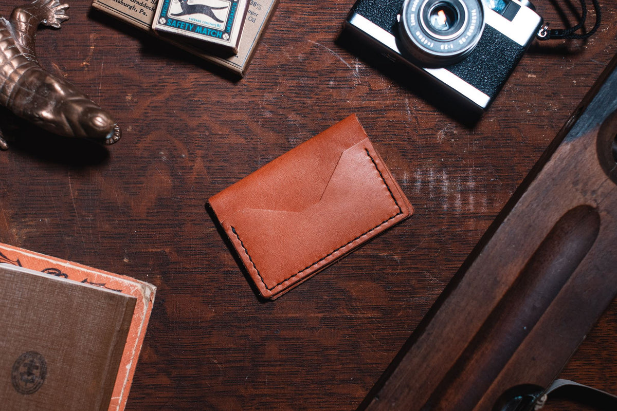 A tan leather bifold card holder wallet with a flap closure, placed on a dark wooden desk surrounded by vintage props including a camera, a brass object, and a book.