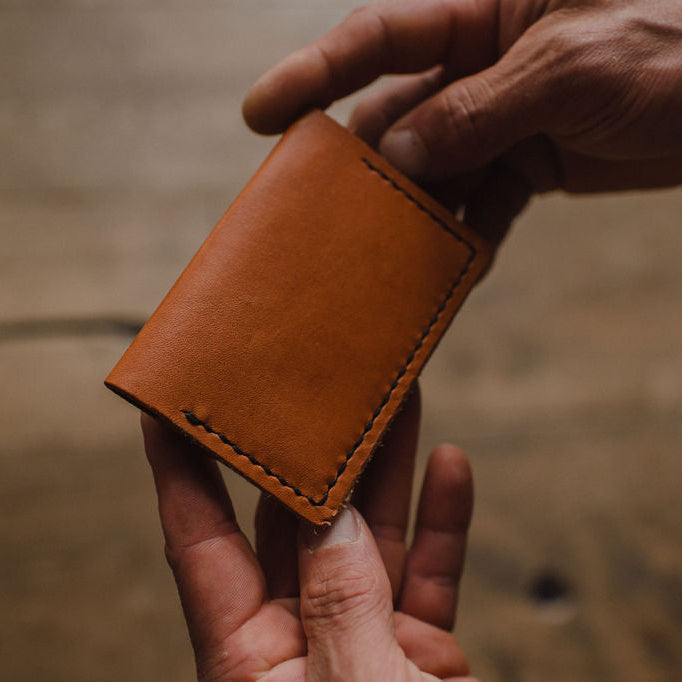 A close-up of a person's hands holding a bifold tan leather card holder wallet against a blurred wooden background.