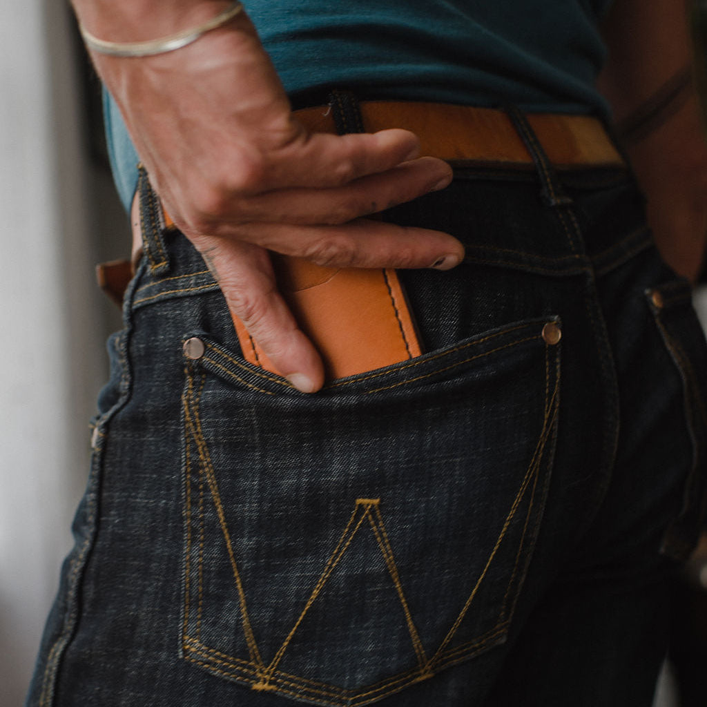 Close-up of a person wearing dark blue denim jeans with a tan leather belt with a tan leather money clip wallet tucked into the back pocket, wearing a teal t-shirt.