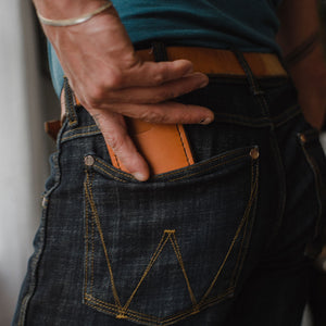 Close-up of a person wearing dark blue denim jeans with a tan leather belt with a tan leather money clip wallet tucked into the back pocket, wearing a teal t-shirt.