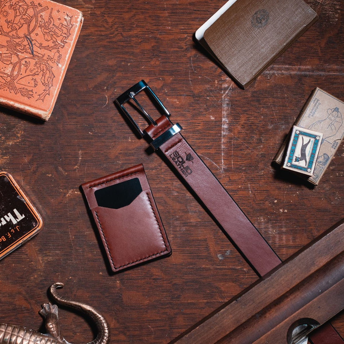 A cognac brown formal leather belt and matching card holder wallet in cognac brown are arranged on a dark wooden tabletop with other styled leather goods and props around them. The belt is brown with a stainless steel buckle and a small embossed logo near the end.