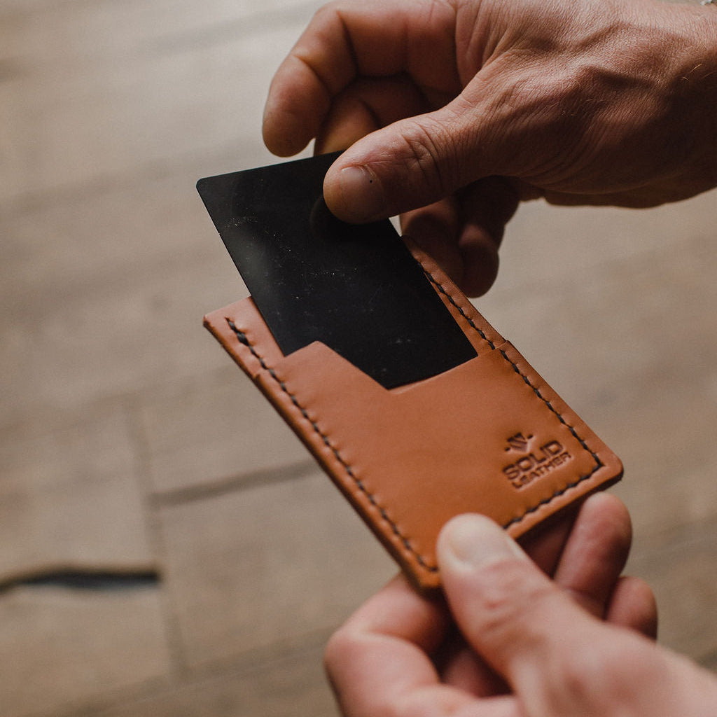 A close-up of a person's hands holding a tan leather card holder with a black card inserted, against a blurred wooden background. The card holder has a debossed logo in the bottom right corner.