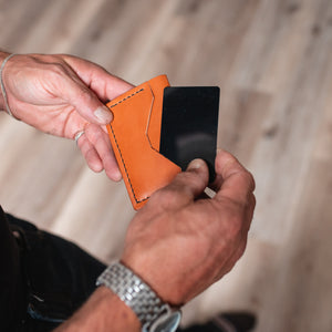 A close-up of a person's hands holding an full-grain tan leather card sleeve with a black card partially inserted, set against a blurred wooden floor background.