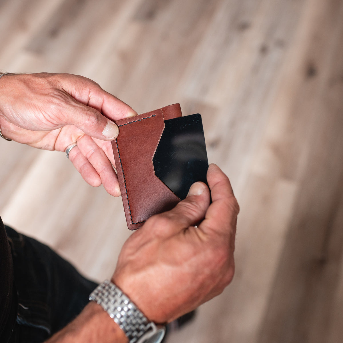 A close-up of a person's hands holding a cognac brown leather card holder wallet from Solid Leather with a black card inserted, set against a blurred wooden floor background.