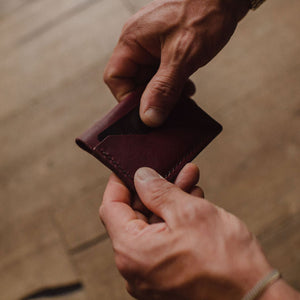 A close-up of a person's hands holding a minimalist burgundy leather bifold card holder wallet against a warm, blurred wooden background.