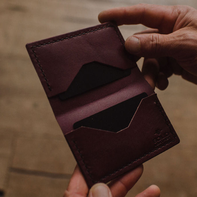 A close-up of a person's hands holding an open burgundy leather bifold cardholder wallet against a warm, blurred wooden background.