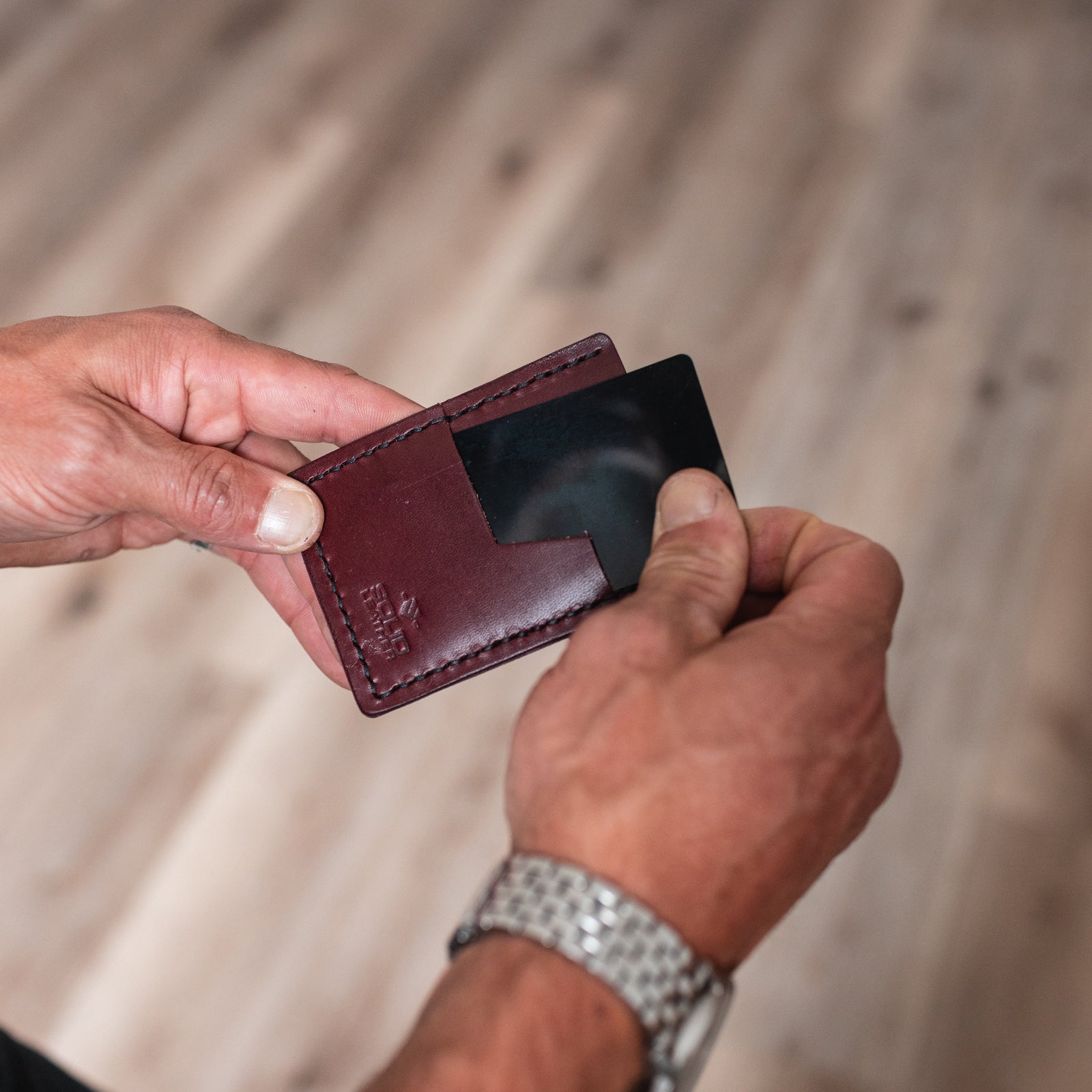 A close-up of a person's hands holding a burgundy leather card holder wallet with a black card inserted, set against a blurred wooden floor background.