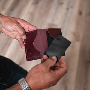 A close-up of a person's hands holding a  burgundy leather card holder wallet with a black card partially inserted, set against a blurred light wood floor background.