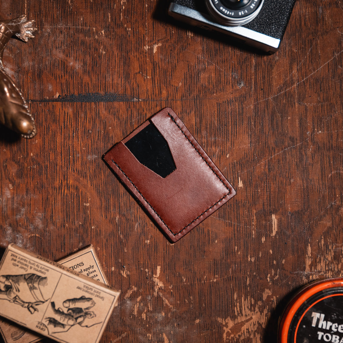A styled flat-lay of a slim leather card holder wallet on a rustic wooden table, surrounded by vintage props including a camera, a lizard figurine, matchboxes, and a tin of tobacco.