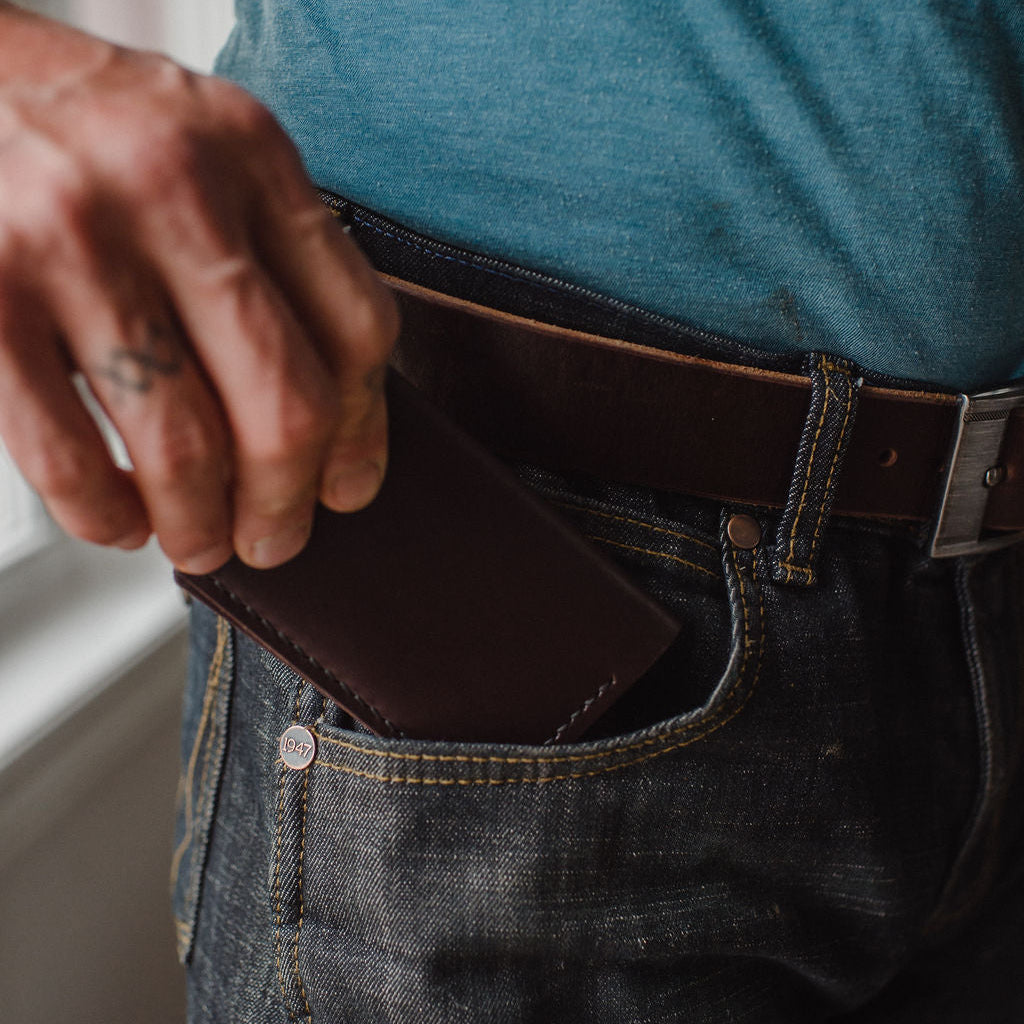 A dark Brown leather bifold cardholder wallet handmade in canada being inserted into the front pocket of dark blue jeans with a matching dark brown Solid Leather belt and blue tshirt 