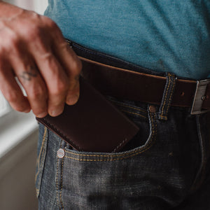 A dark Brown leather bifold cardholder wallet handmade in canada being inserted into the front pocket of dark blue jeans with a matching dark brown Solid Leather belt and blue tshirt 