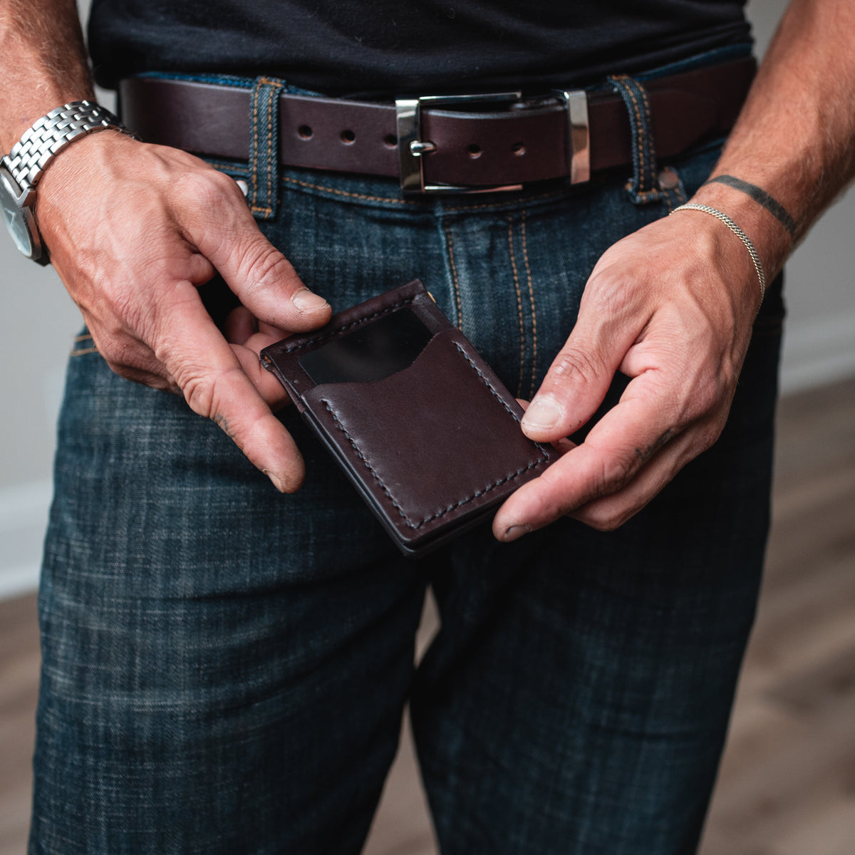 a mans hands holding a dark brown leather money clip bifold cardholder wallet with matching full-grain leather belt being worn on blue jeans 