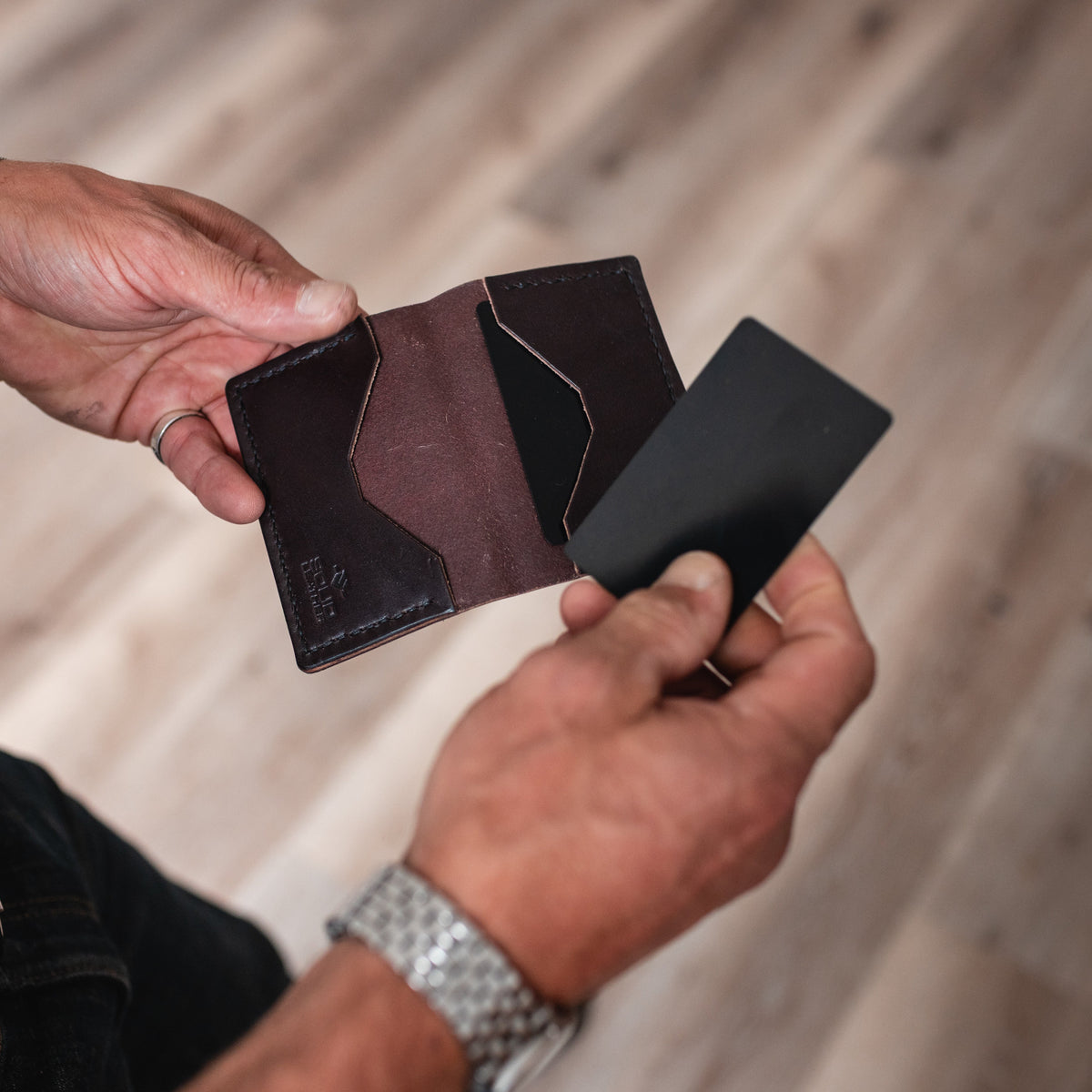 A close-up of a person's hands holding an open dark brown leather cardholder wallet and a black card, with a blurred wooden floor in the background.