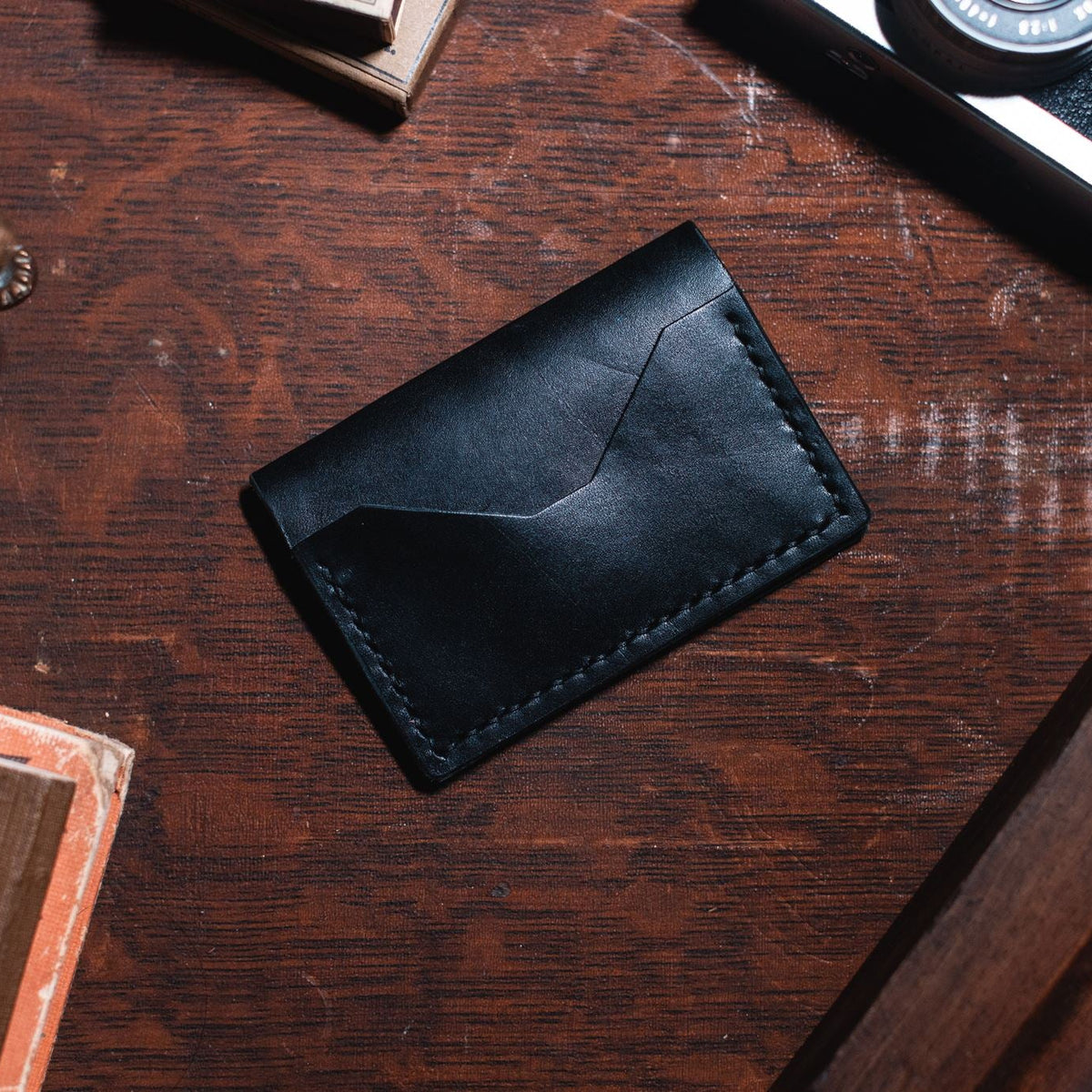 A black leather bifold card holder wallet with visible stitching, placed on a dark wooden desk surrounded by vintage props including a camera, a brass figurine, and a book.