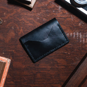 A black leather bifold card holder wallet with visible stitching, placed on a dark wooden desk surrounded by vintage props including a camera, a brass figurine, and a book.