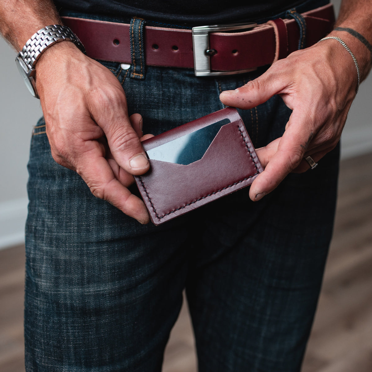 A close-up of a man's hands holding a burgundy leather card holder wallet, wearing dark blue jeans and a matching burgundy  leather belt.