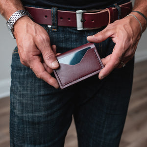 A close-up of a man's hands holding a burgundy leather card holder wallet, wearing dark blue jeans and a matching burgundy  leather belt.