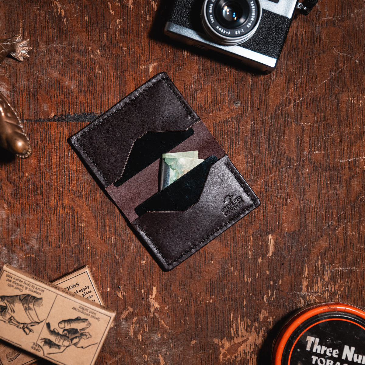 A dark brown leather bifold wallet with a cardholder and a visible cash compartment, styled on a rustic wooden tabletop with a camera, camera lens, lizard figurine, stamps, and a round tin nearby.