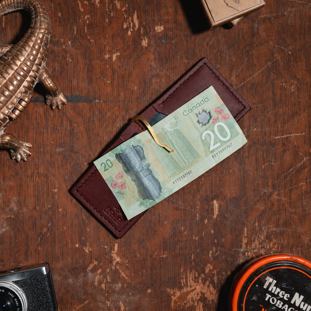 A cognac brown, full-grain  leather bi-fold money clip wallet holding Canadian twenty-dollar banknotes, arranged on a rustic wooden tabletop alongside a bronze alligator figurine, a vintage camera, and a tobacco tin.