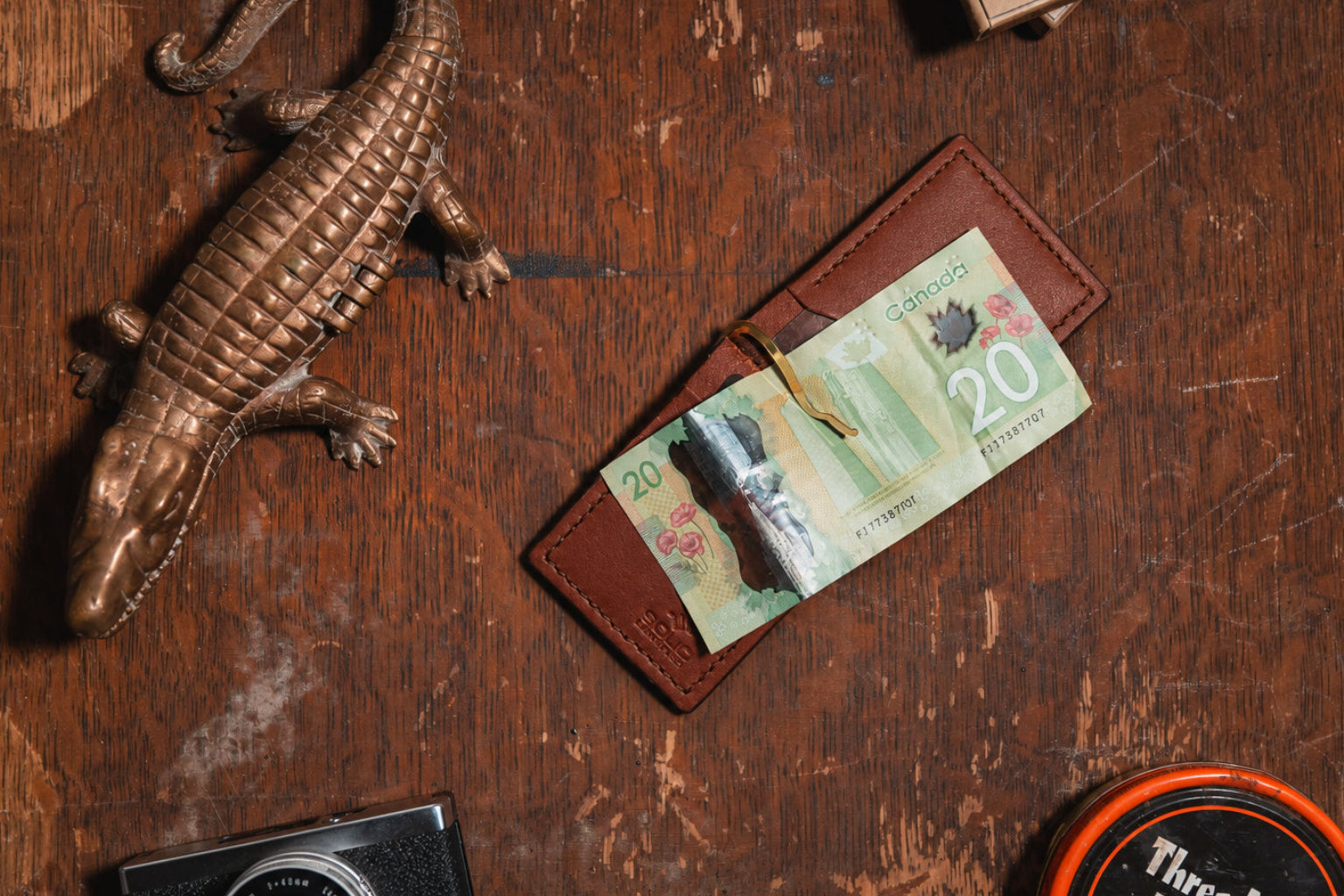 an open cognac brown leather money clip wallet with $20 Canadian banknote on a wooden table with vintage props 