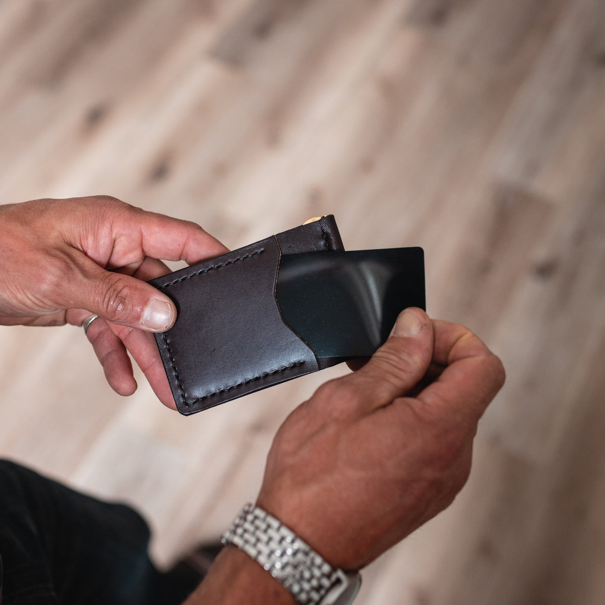 A close-up of a person's hands holding a slim dark brown leather money clip card holder wallet with a black card partially inserted, set against a blurred wooden floor background.
