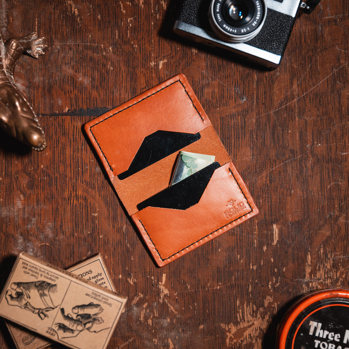 A tan leather card holder wallet displayed in a flat-lay arrangement on a rustic wooden table alongside a camera, a lizard figurine, and other vintage-style props.