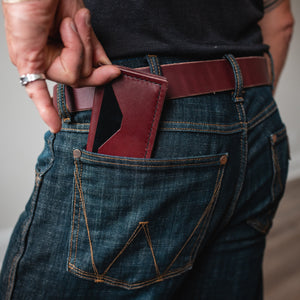 A close-up lifestyle shot of a person wearing dark blue denim jeans and a burgundy full-grain leather belt, showing a burgundy leather card holder wallet tucked into the back pocket.