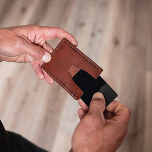 A close-up of a person's hands holding a cognac brown leather card holder with a black card inserted, set against a blurred wooden floor background.