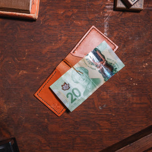 A tan leather cardholder wallet holding a Canadian 20-dollar banknote, placed on a dark wooden desk with vintage props. handmade by jesse bateson of Solid Leather in Peterborough Ontario Canada 