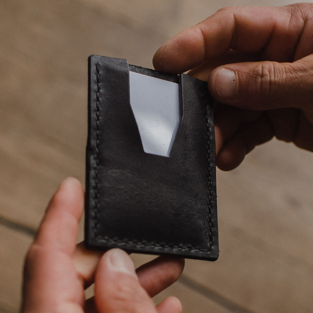 A close-up of a person's hands holding a slim black leather card holder wallet with a clear ID window, set against a blurred wooden background.