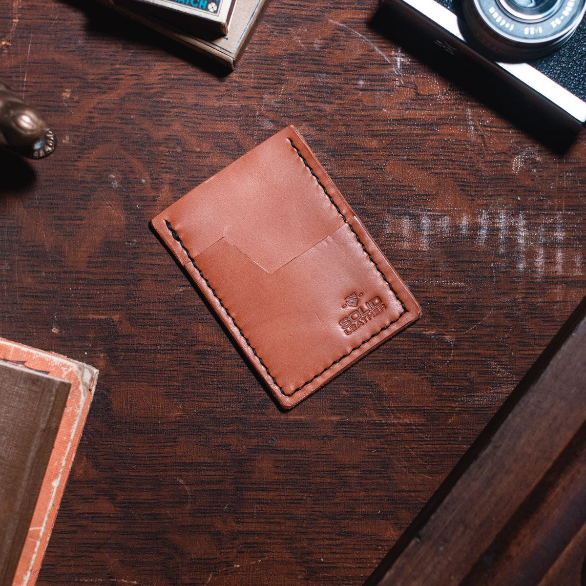 A tan leather card holder wallet with a debossed logo in the bottom right corner, placed on a dark wooden desk surrounded by vintage props like a camera, a book, and a metal figurine.