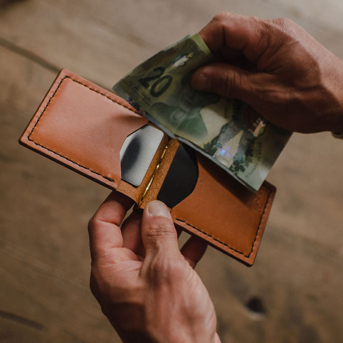 A close-up of a tan leather money clip  wallet being opened, with a person's hands inserting an Canadian twenty-dollar note. The person is wearing a silver link watch, and the background is a warm, out-of-focus wooden surface.