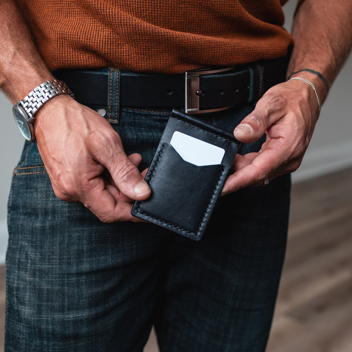 A close-up of a man's hands holding a slim black leather money clip card holder wallet, wearing a rust-orange knit sweater, matching black leather belt, and dark blue jeans in an indoor setting with a wood floor.
