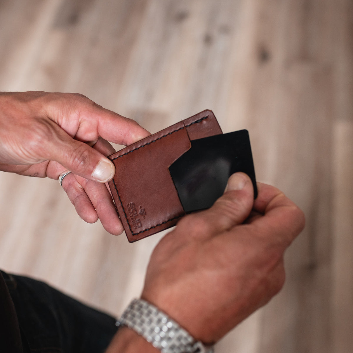 A close-up of a person's hands holding a cognac brown leather cardholder wallet and sliding a black card into it, set against a blurred wooden floor background.