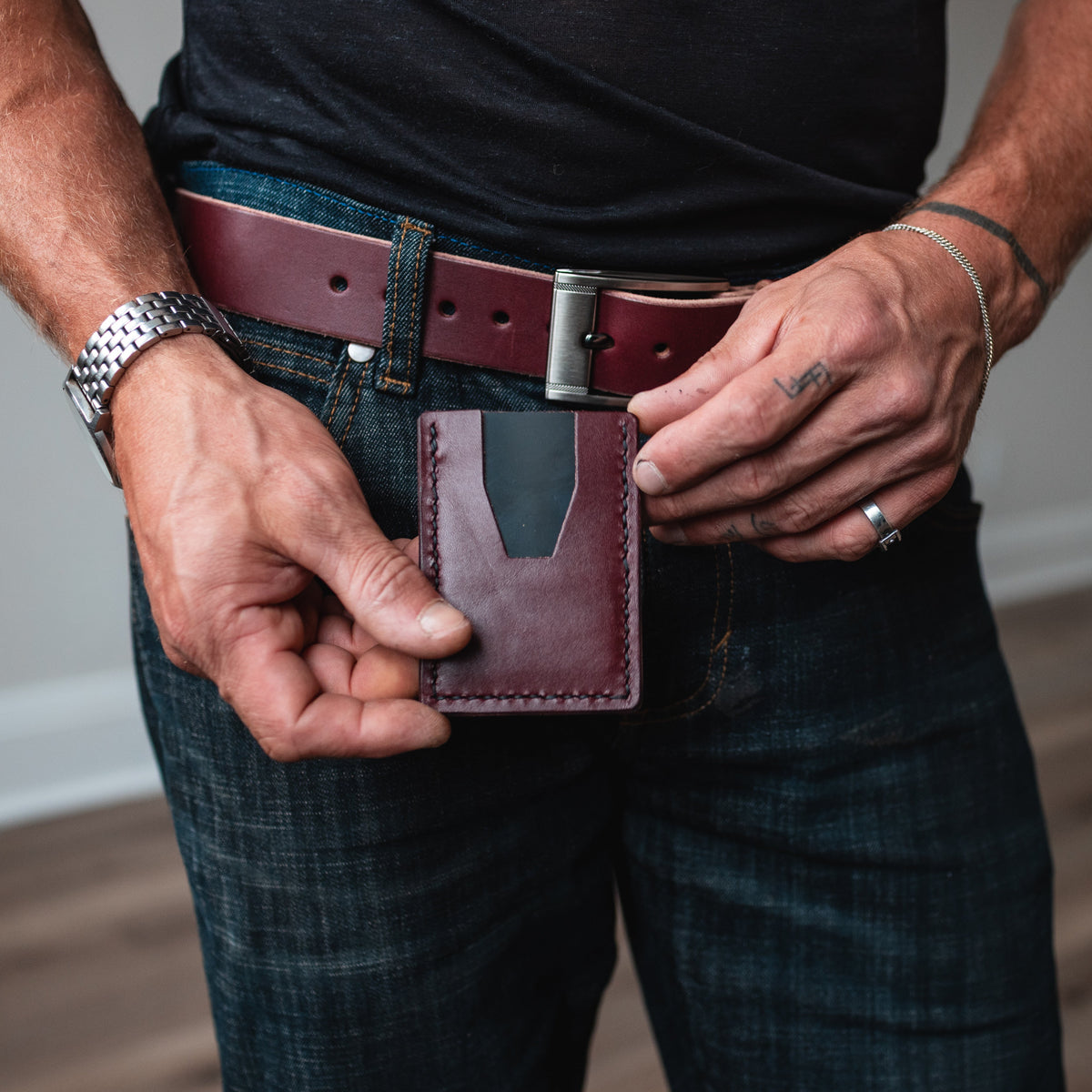 A close-up of a man's hands holding a burgundy leather card holder wallet, with his matching full grain burgundy leather belt and blue jeans in an indoor setting with a wood floor.
