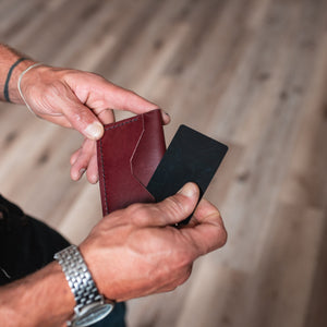 A close-up of a person's hands holding a dark burgundy leather card holder wallet and a black card, set against a blurred wooden floor background.