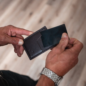 A close-up of a person's hands holding an open dark brown leather cardholder wallet, showing its interior slots, against a blurred light wood floor background.