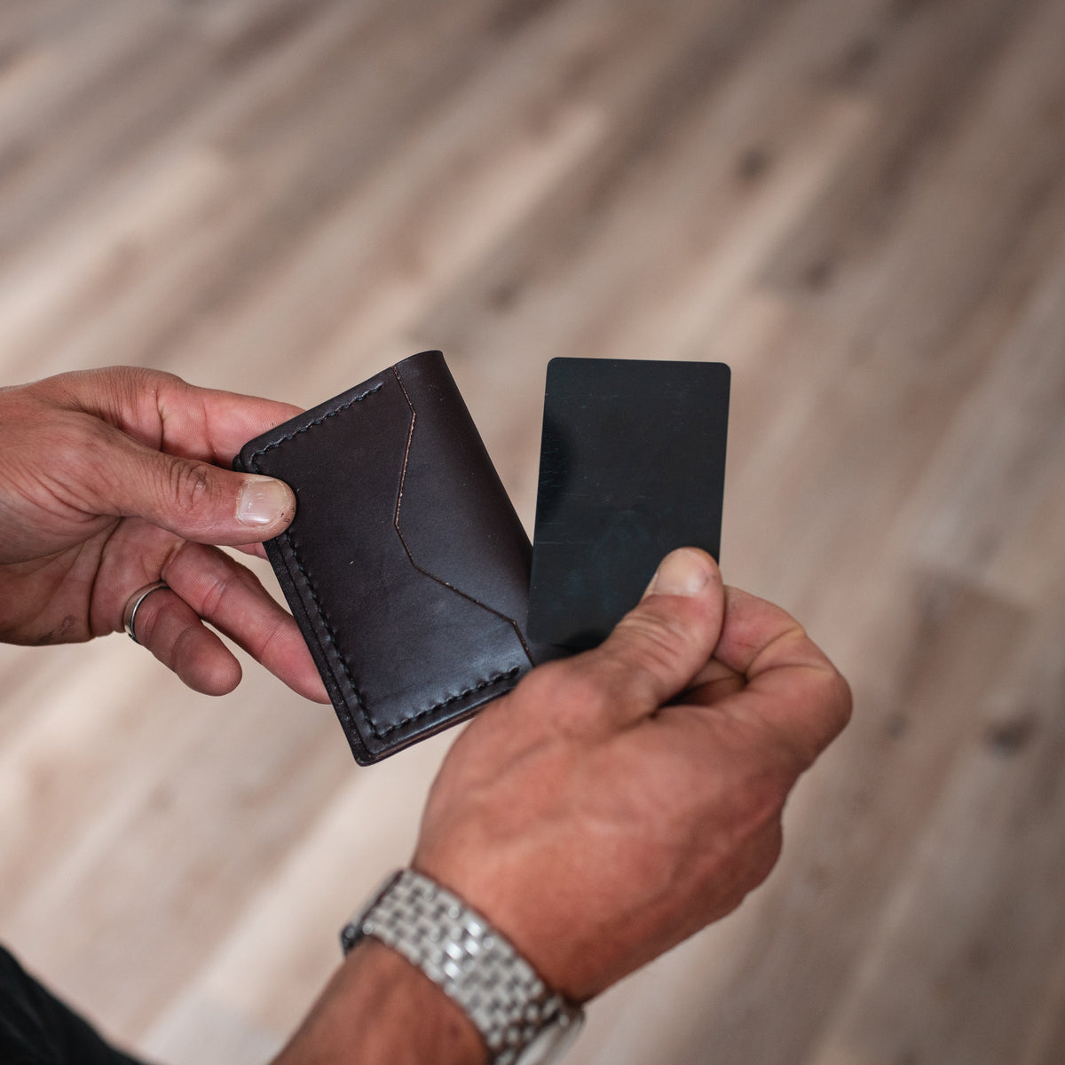 A close-up of a person's hands holding a dark brown leather card holder wallet next to a black card, with a blurred wooden floor in the background.