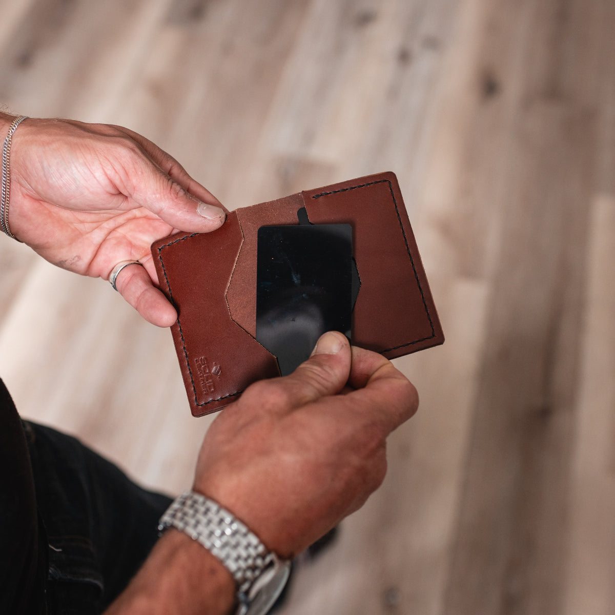 A close-up of a person's hands holding an open cognac brown leather cardholder wallet with a black card inside, set against a blurred light wood plank floor background.