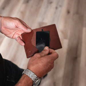 A close-up of a person's hands holding an open cognac brown leather cardholder wallet with a black card inside, set against a blurred light wood plank floor background.