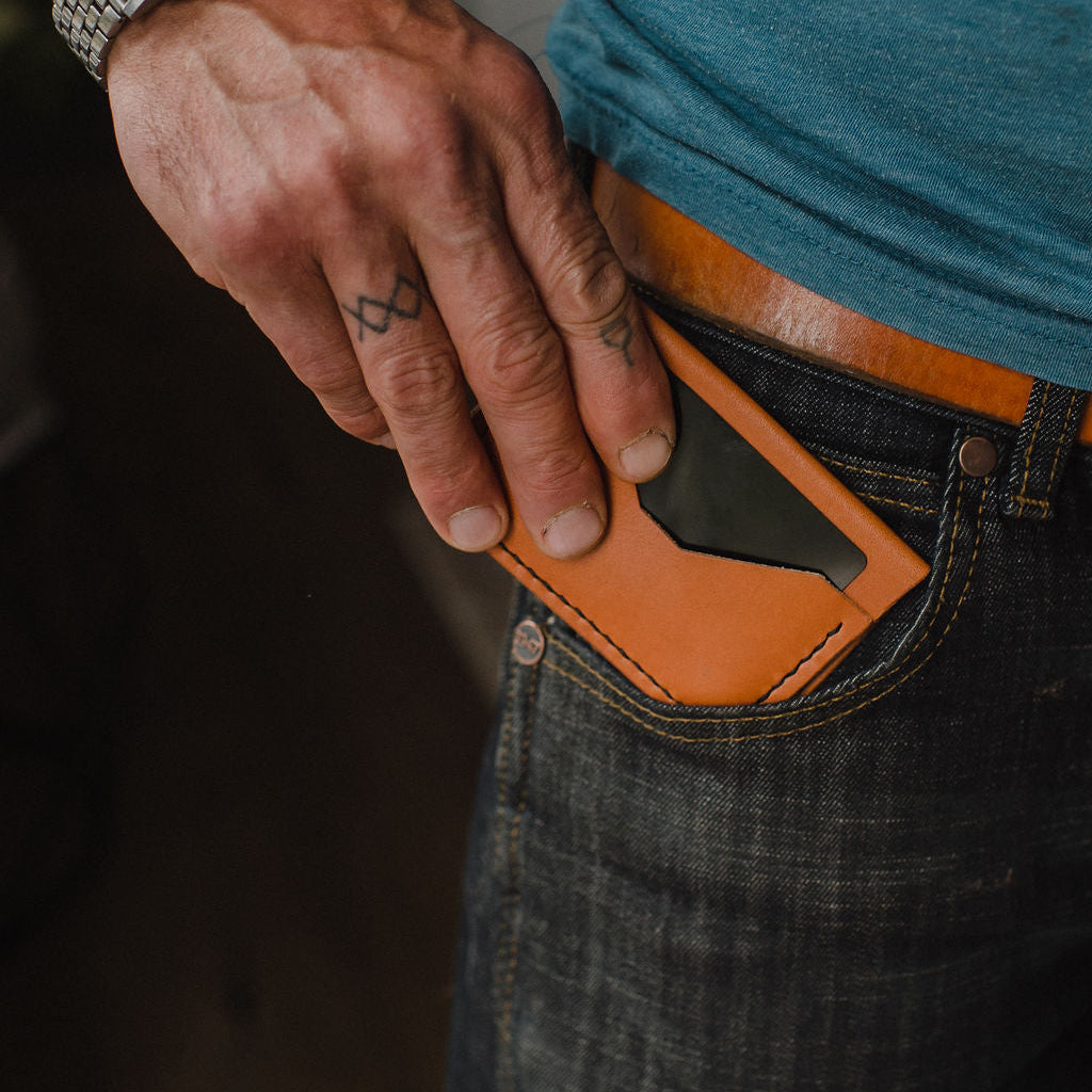 A close-up of a man's hand inserting a  tan solid leather bifold card holder wallet into the front pocket of his dark blue denim jeans, secured by a matching Solid Leather tan belt.