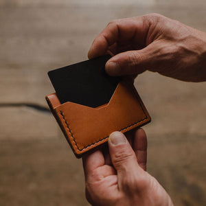 A close-up of a person's hands holding a Solid Leather slim, tan leather bifold card holder wallet with a black card partially inserted, set against a blurred wooden background.