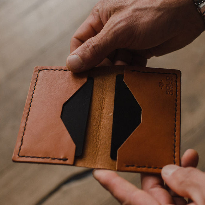 A close-up of a person holding an open tan leather bifold card holder wallet with black interior lining, showing its card slots and stitching, against a blurred wooden background. handmade in Peterborough Ontario by jesse bateson of Solid Leather 