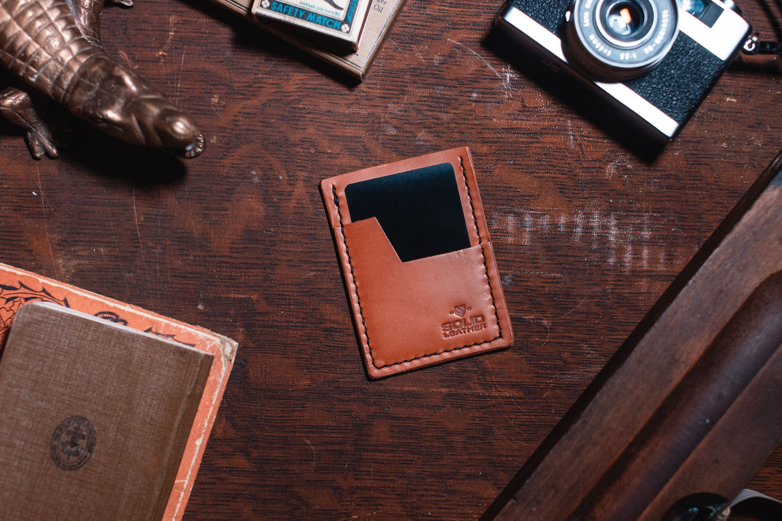 A tan leather card holder wallet with a black card slot, placed on a dark wooden desk surrounded by vintage props including a camera, a small figurine, and an old book.